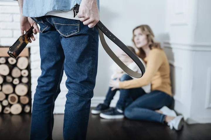 woman and child cowering in corner and man standing with a belt