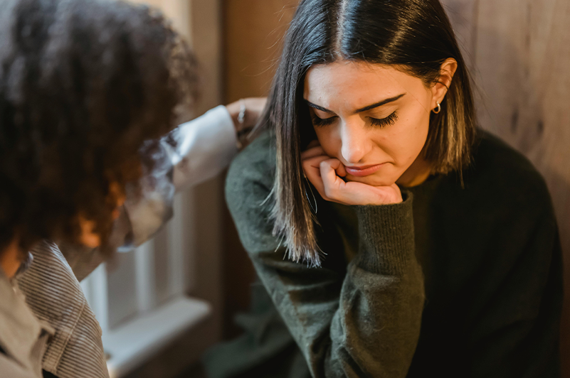 woman looking down and another woman with her hand on her shoulder